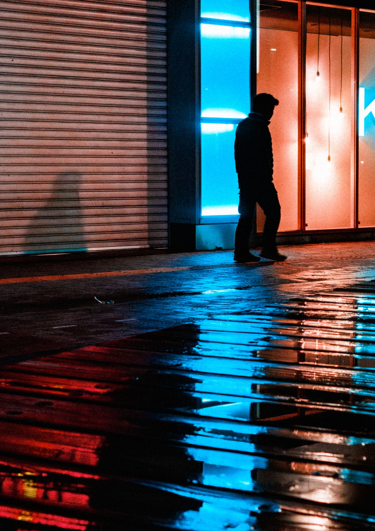 A silhouetted figure walking between blue and orange storefront lights reflected on wet pavement.